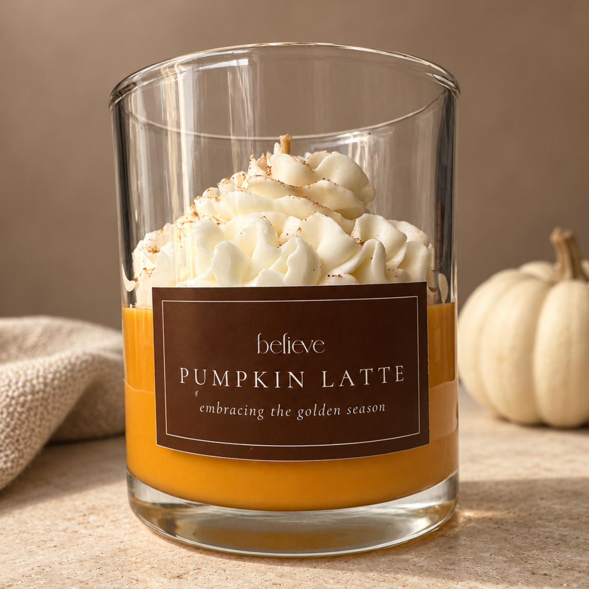 Pumpkin Latte candle in a clear glass jar, its amber wax topped with whipped cream-style wax, beside a white pumpkin and a knitted throw