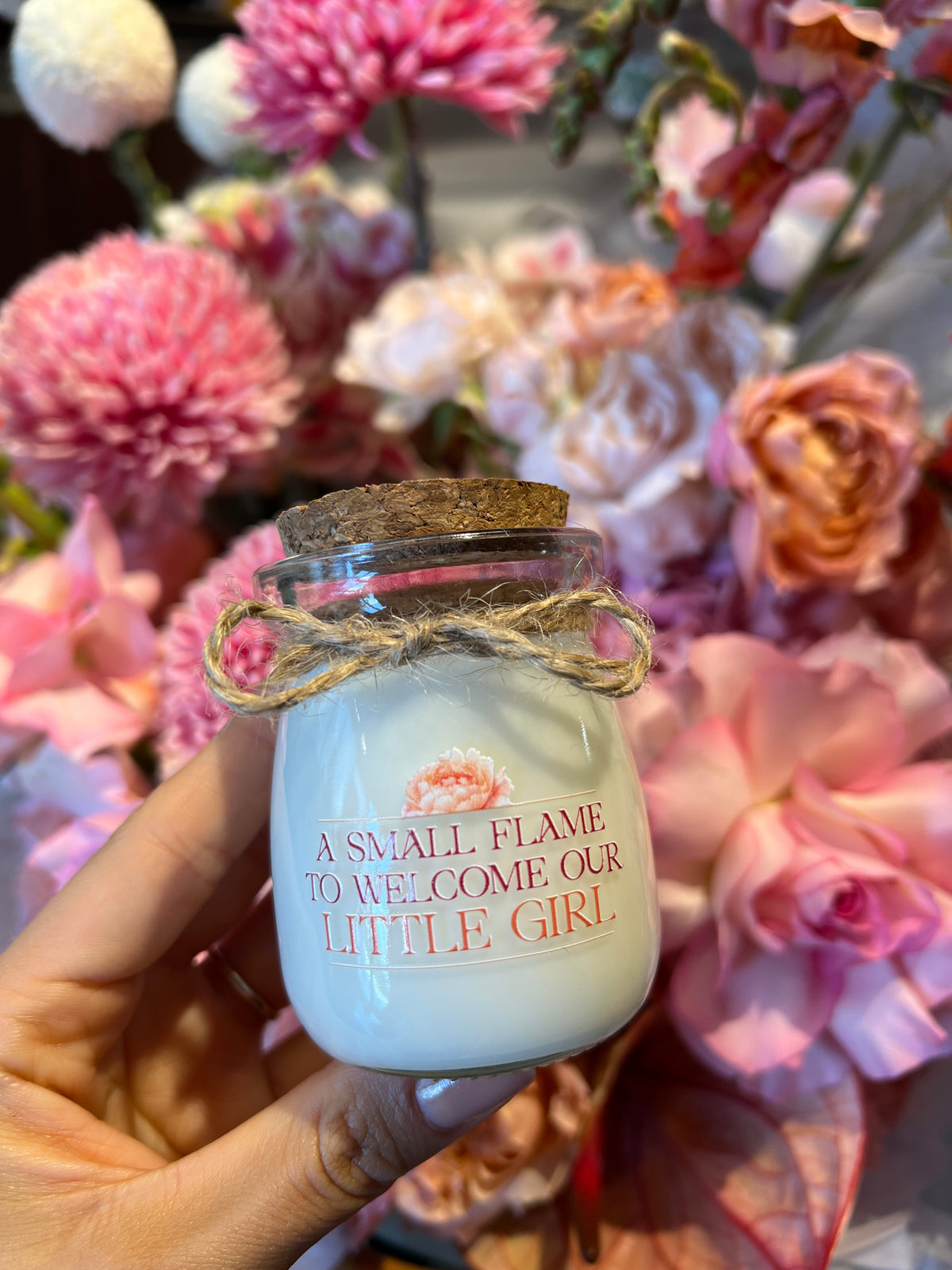 Hand holding a personalised BELIEVE candle with a custom printed label, in front of fresh flowers